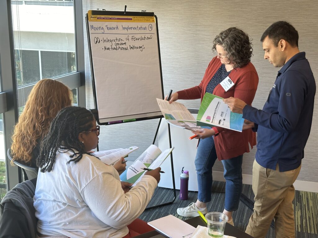 Four standards writers refer to the Reimagining CS Pathways report as they write ideas down on a post-it board.
